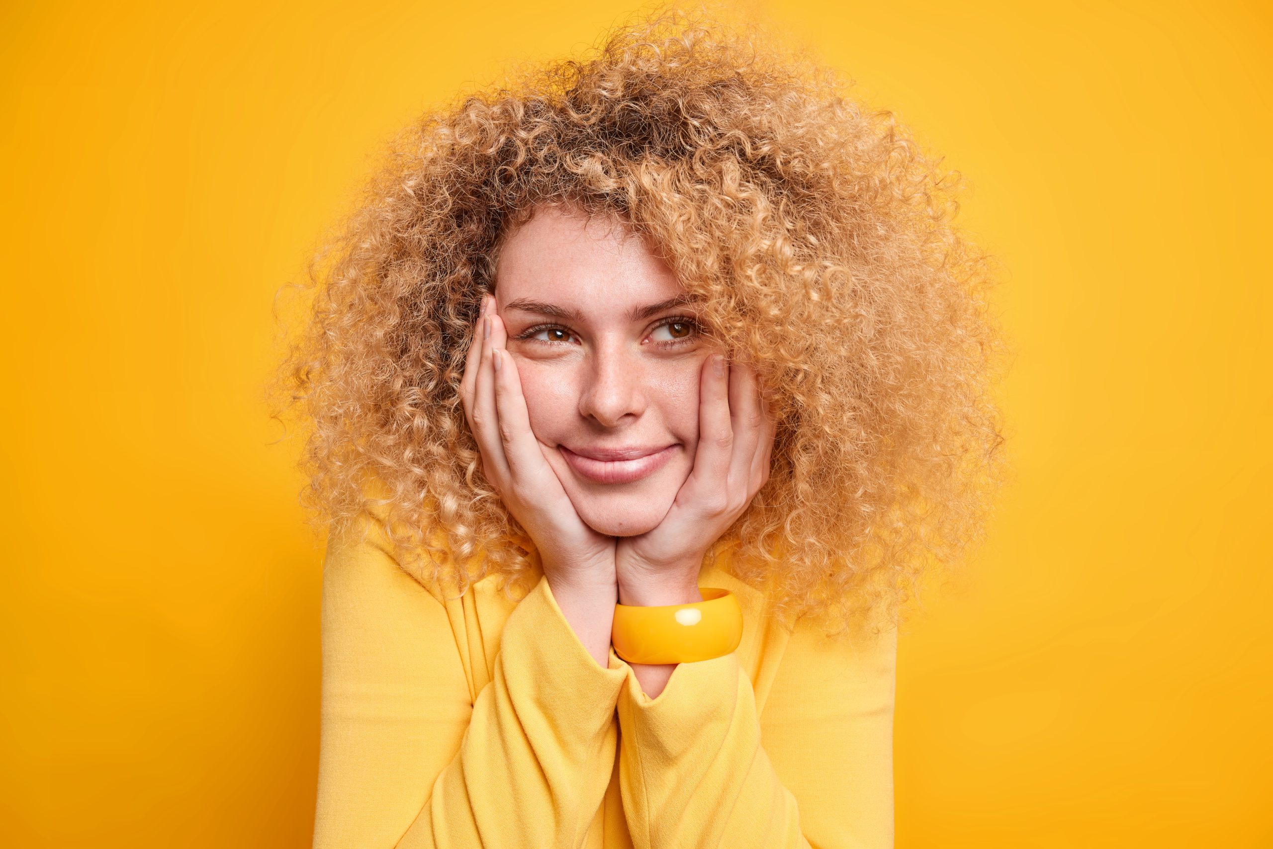 Headshot of pretty beautiful young European woman has dreamy expression keeps hands on cheeks thinks about something pleasant dressed formally looks away isolated over vivid yellow background.